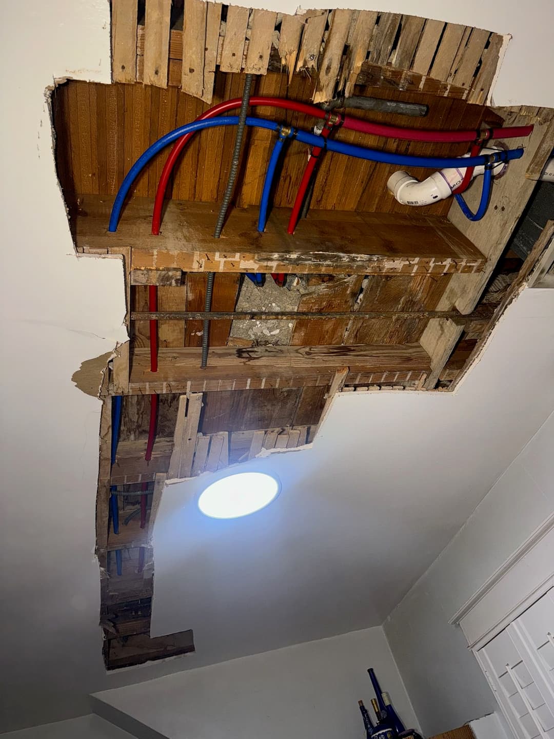 Damaged ceiling revealing wooden beams and exposed plumbing with blue and red pipes.