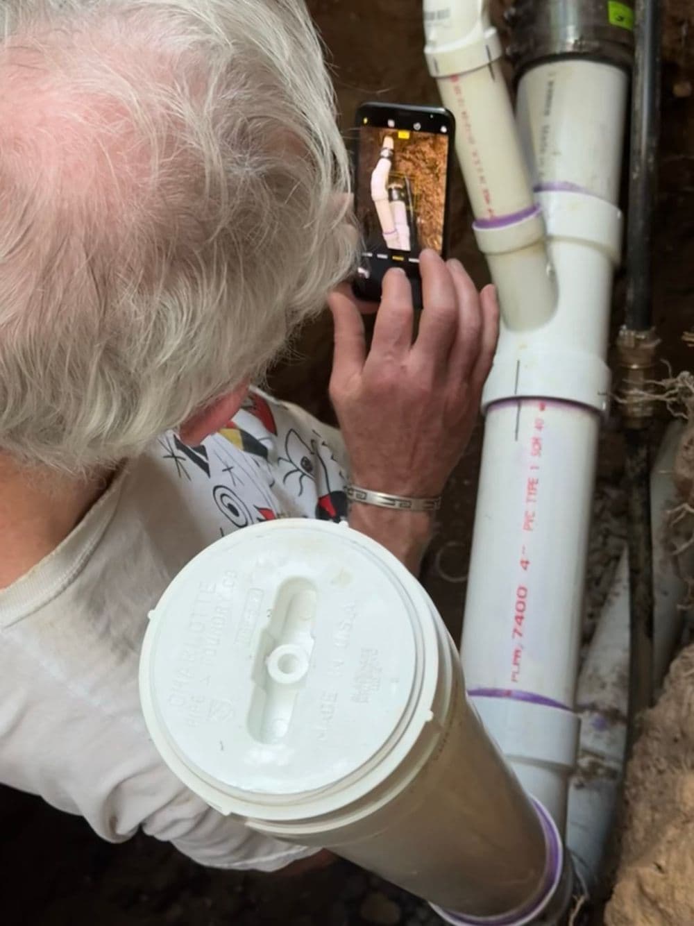 Person photographing a PVC pipe installation in a trench with smartphone.