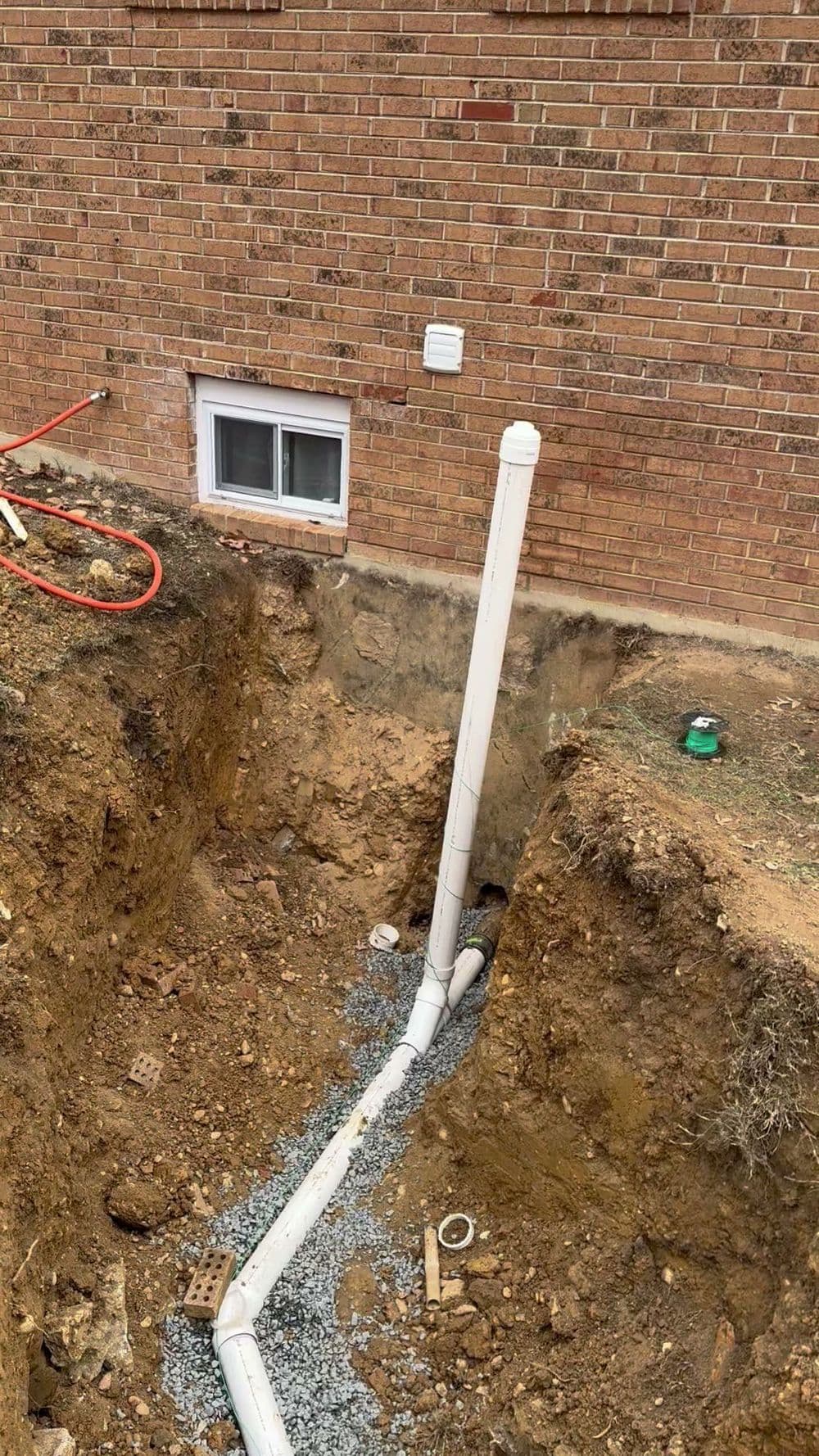 Excavated trench with white drainage pipes near a brick foundation and window installation site.