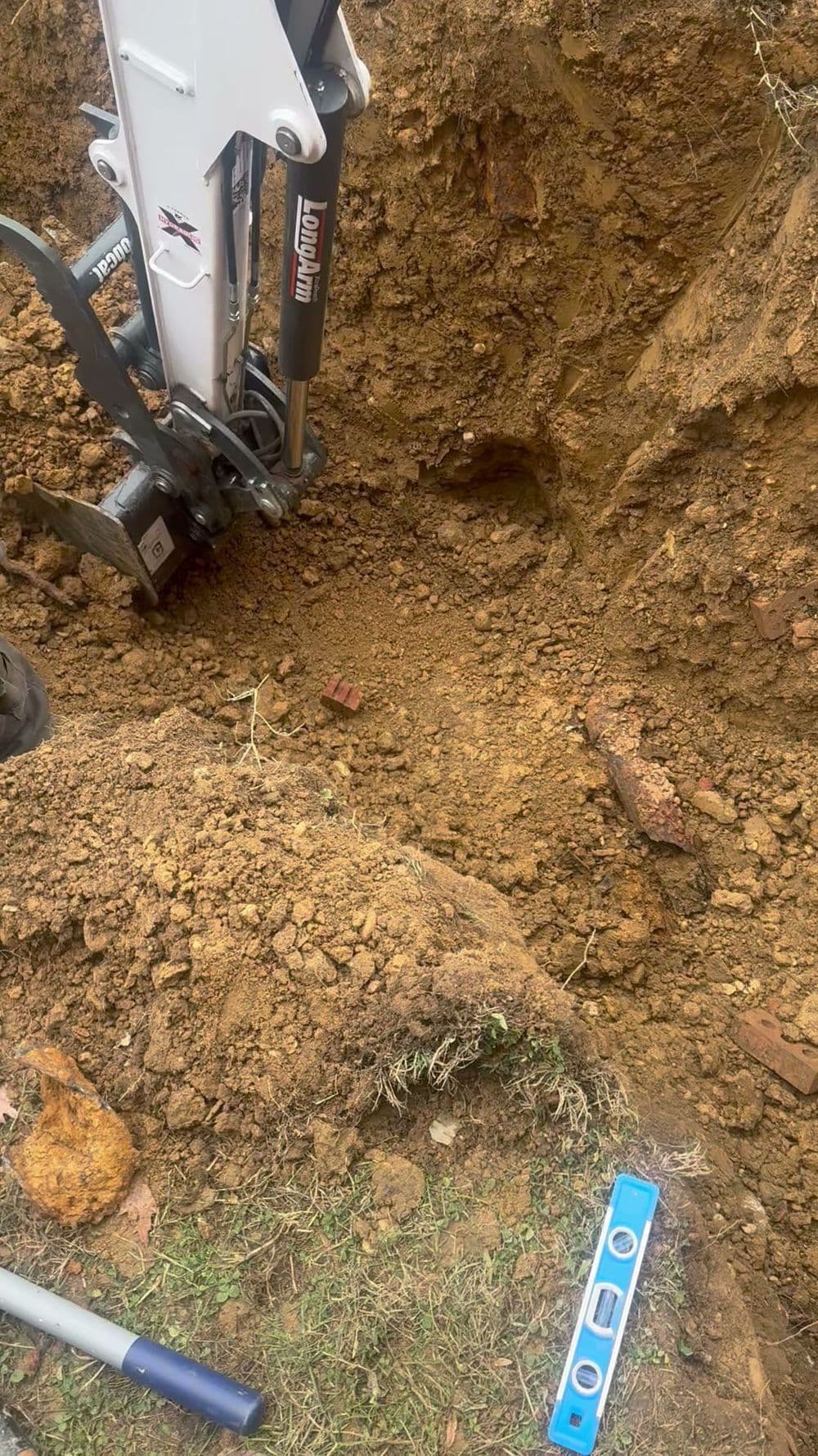 Excavator digging in soil with a level tool and bricks visible in the excavation site.