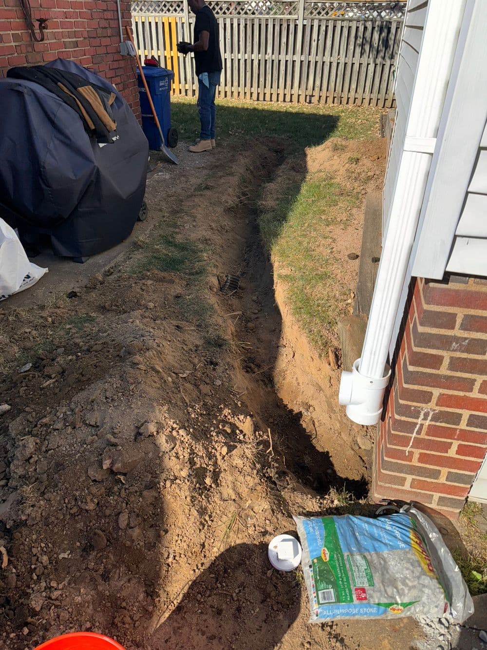 Person digging a trench in the yard, with gardening supplies nearby and a fence in background.