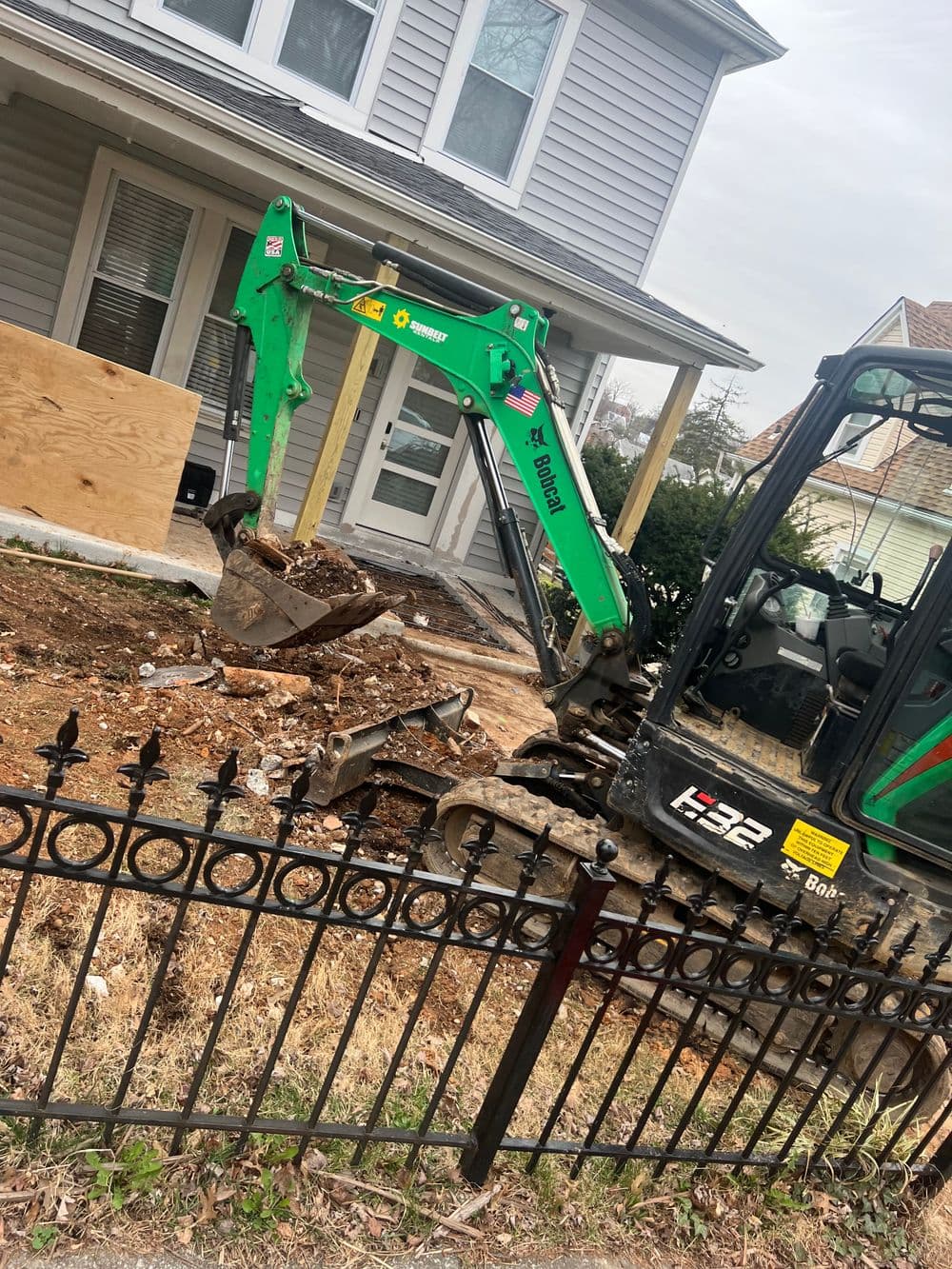 Excavator digging foundation near residential home with wooden boards and fence.
