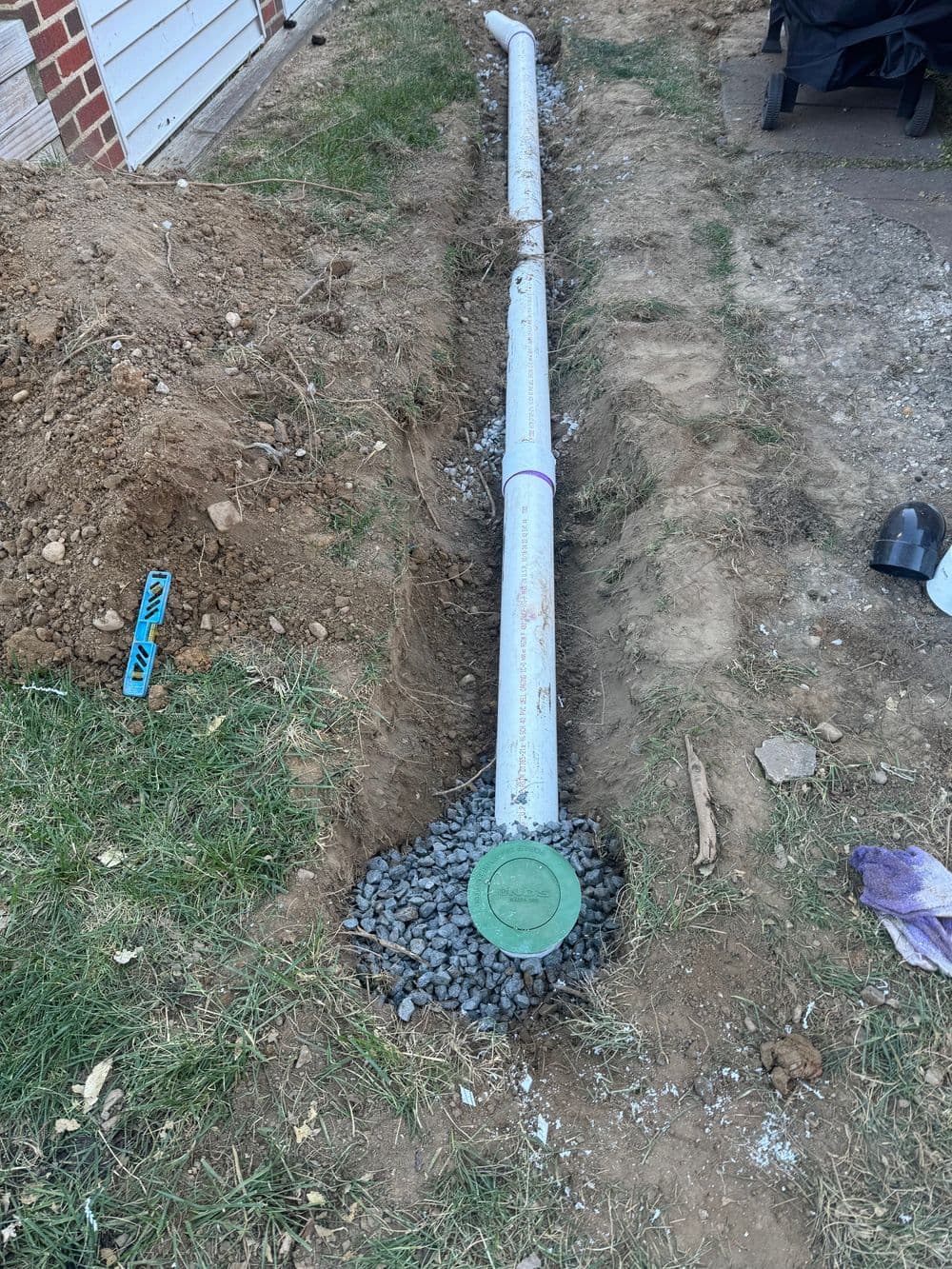 Installed drainage pipe laid in a trench with gravel and access cover, surrounded by dirt and grass.