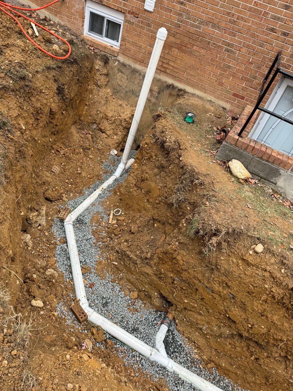 Trenched site showing new white PVC piping installation with gravel base near brick building.