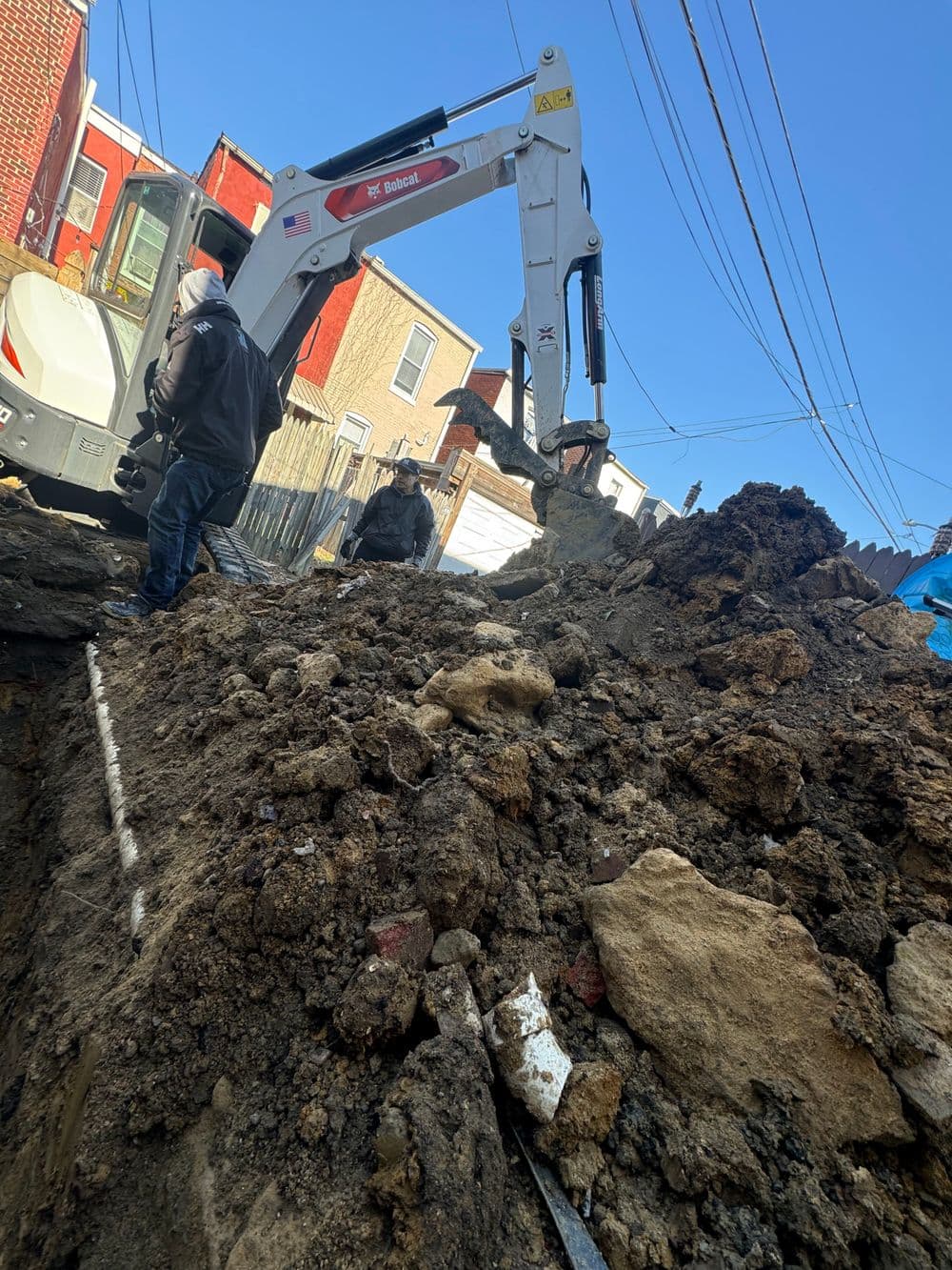 Construction workers operating a backhoe in a residential area with dirt mound and blue sky.
