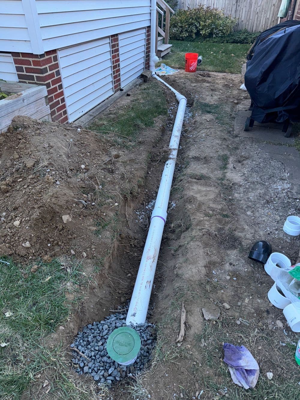 Drainage pipe installation in a trench beside a house, with gravel and tools nearby.