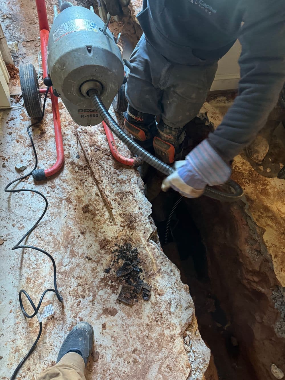 Plumber using a drain snake to clear a deep trench in a residential floor.
