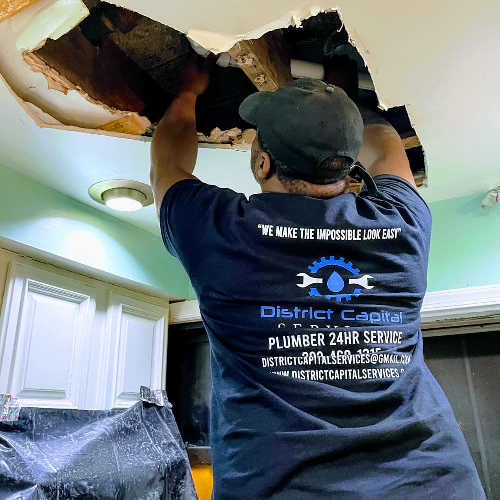 Plumber repairing a ceiling leak in a kitchen wearing District Capital Services shirt.