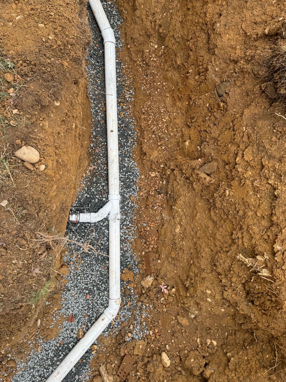 Trenched area showing white drainage pipe installed over gravel in soil.