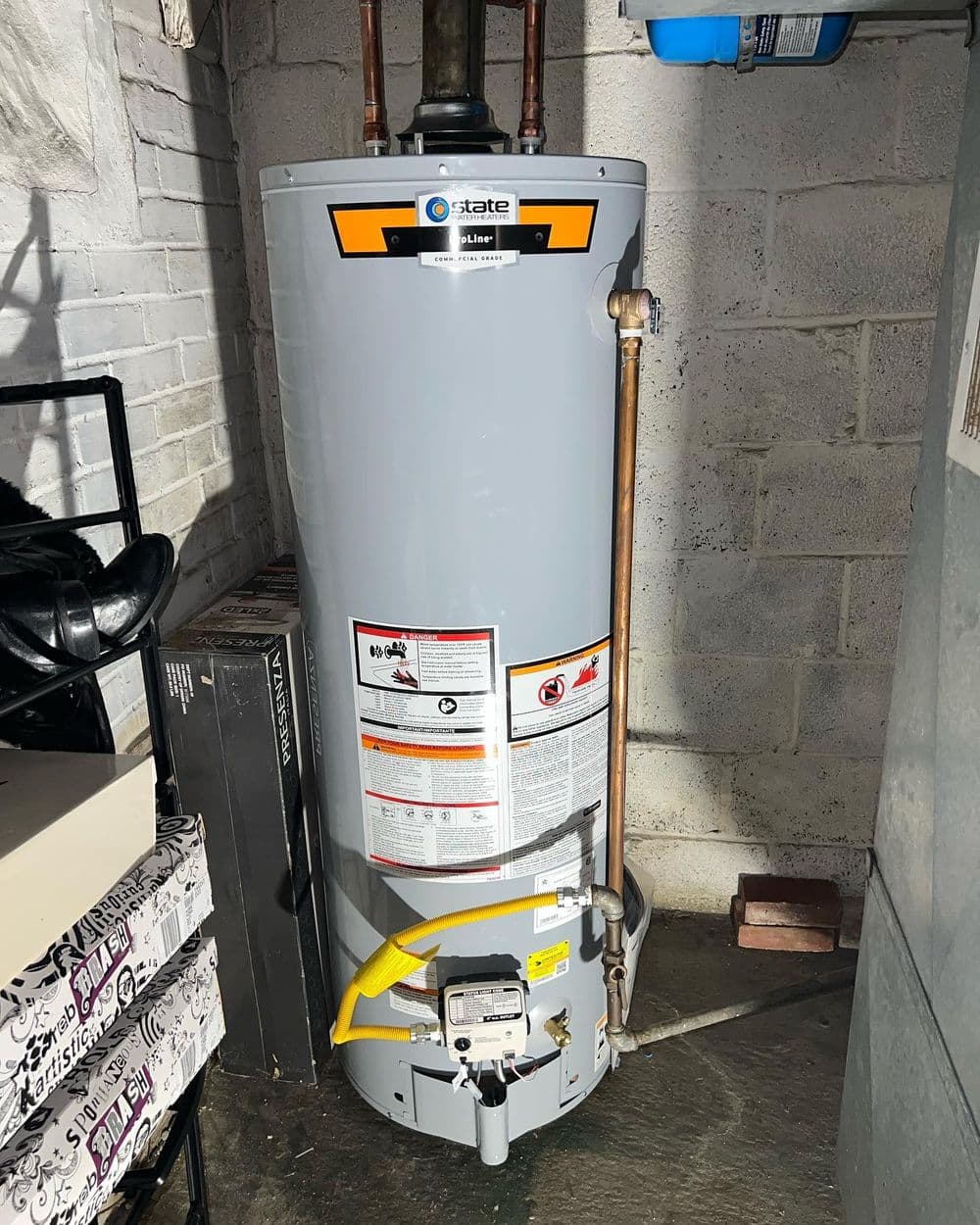 State water heater in a basement with pipes and safety labels visible.